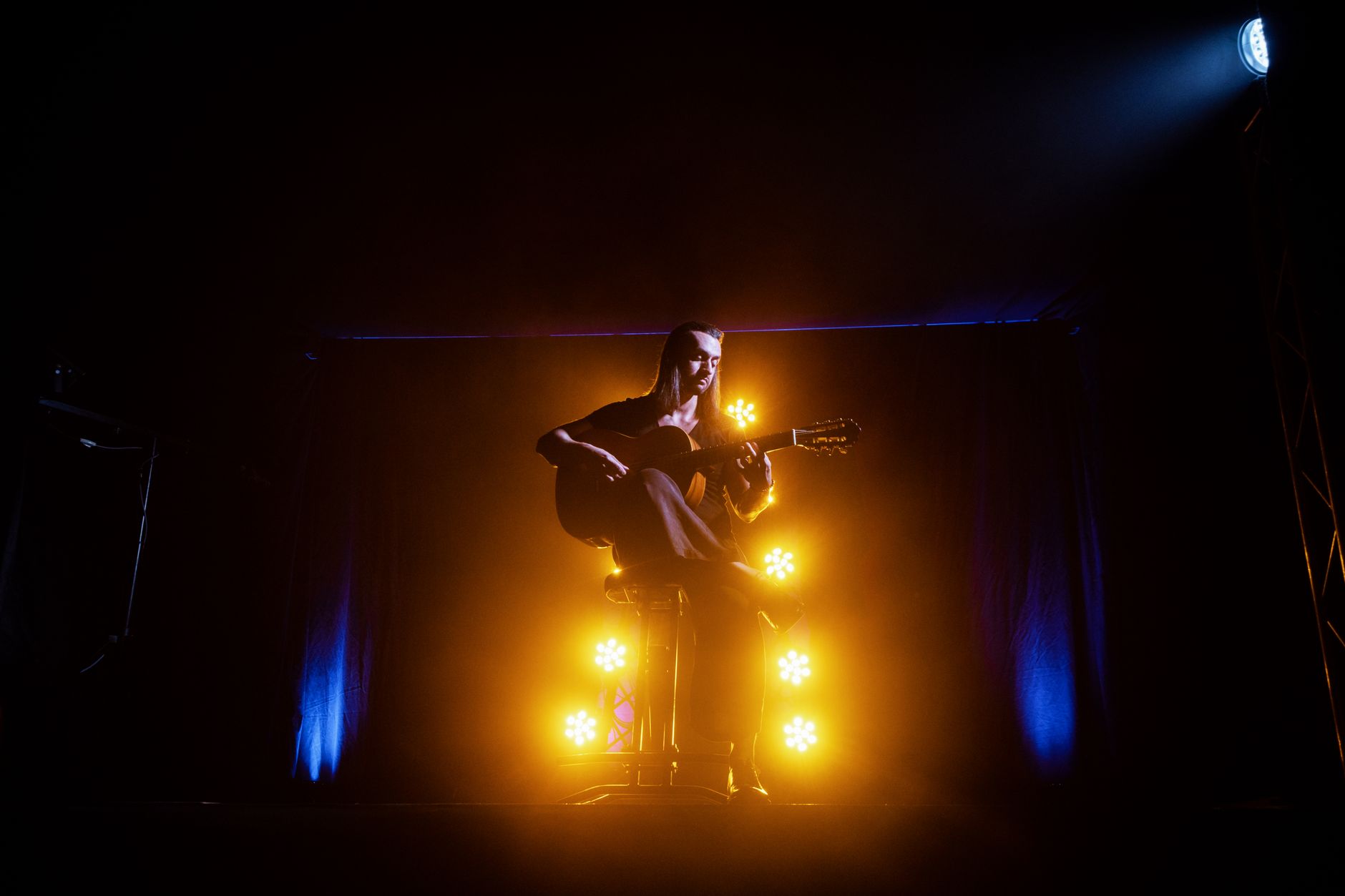 Un músico toca una guitarra acústica en un escenario, iluminado por luces dramáticas amarillas y azules.