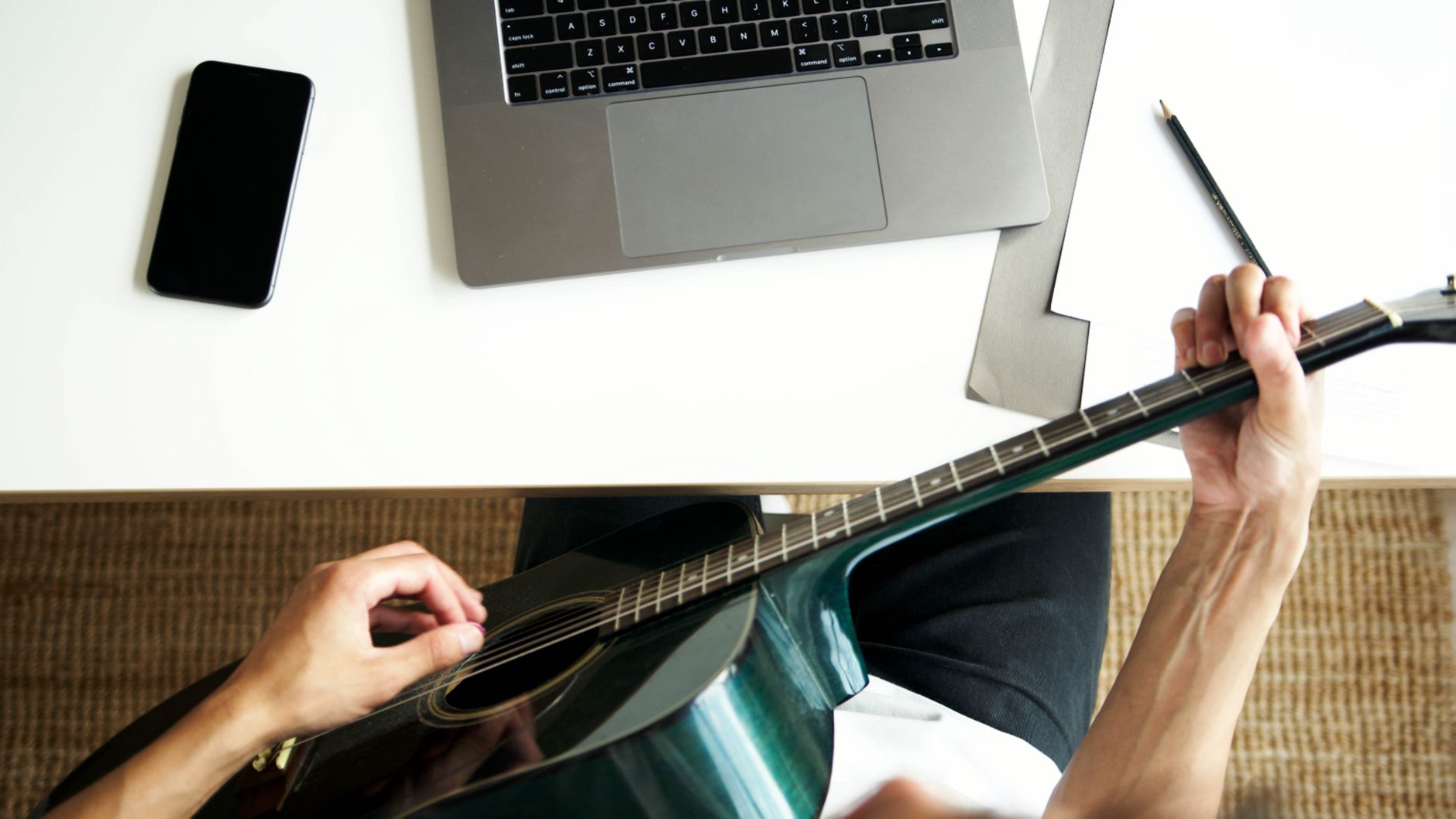 Guitarist composing music at a desk with laptop, paper, and acoustic guitar.