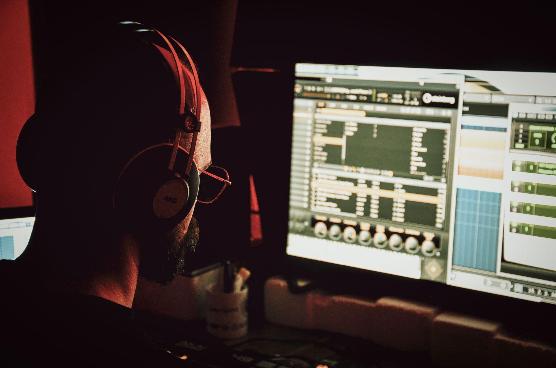 Vista posterior de un freelancer anónimo con barba y auriculares trabajando en la computadora con un programa profesional y escuchando pistas de música por la tarde en su apartamento