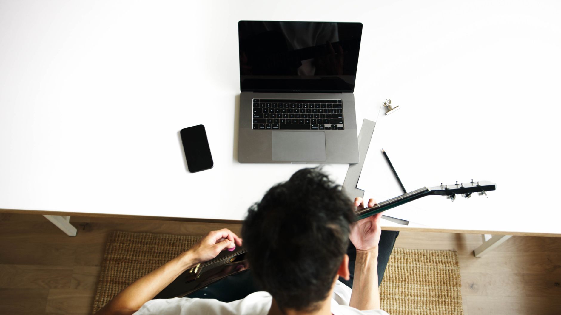Overhead shot of a man playing guitar next to a laptop and smartphone on a desk.
